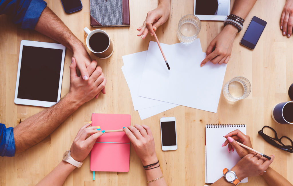 High angle view of group of people sitting at the conference table, discussing, brainstorming. Digital tablets, smart phones, notebooks, coffees on the table. Unrecognizable people.