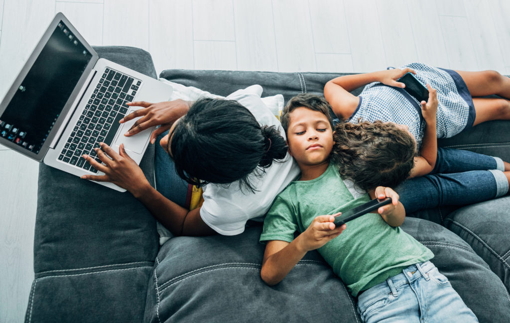 Mixed race family sitting on the sofa at home and using digital devices. African-american mother using laptop while her two children using smartphones.