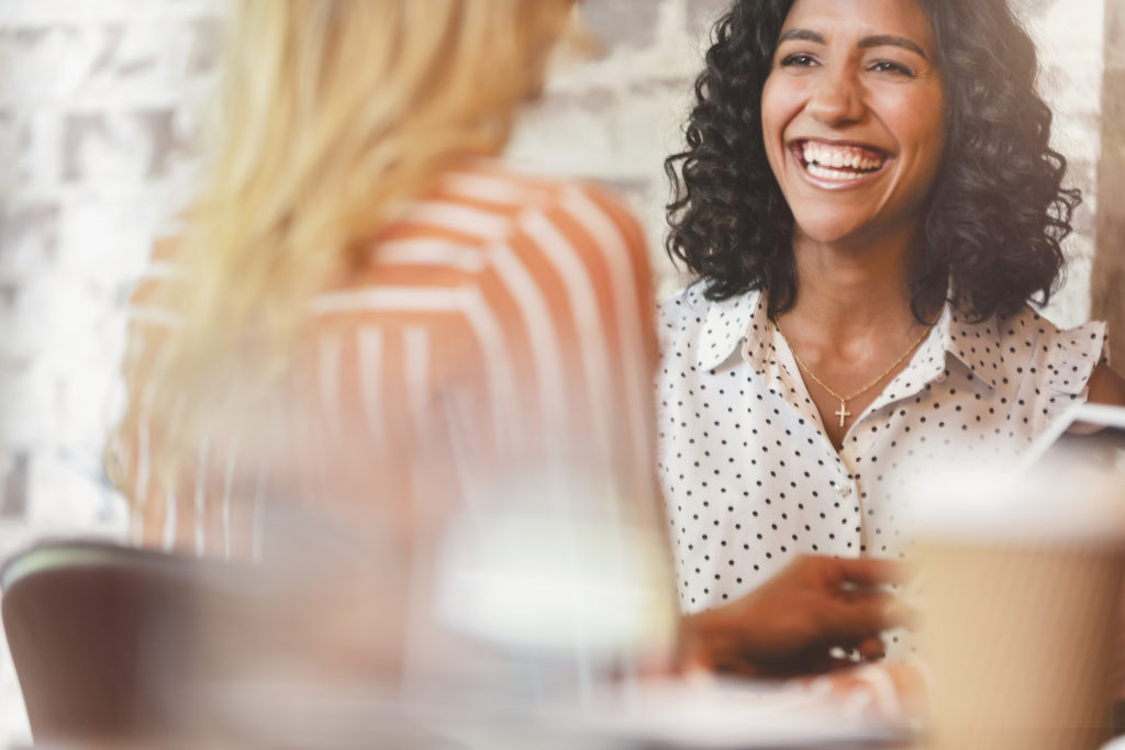 Business colleagues having a conversation. They are both young business people casually dressed in a modern office. Could be an interview or consultant working with a client. She is listening and smiling. One person has her back to us. Mixed ethnic group. One is African American and the other is Caucasian