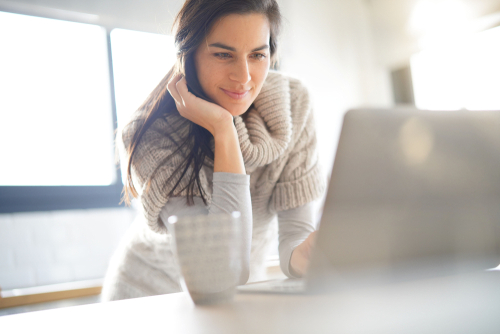 woman reading on laptop on table with coffee mug and lots of light coming in windows