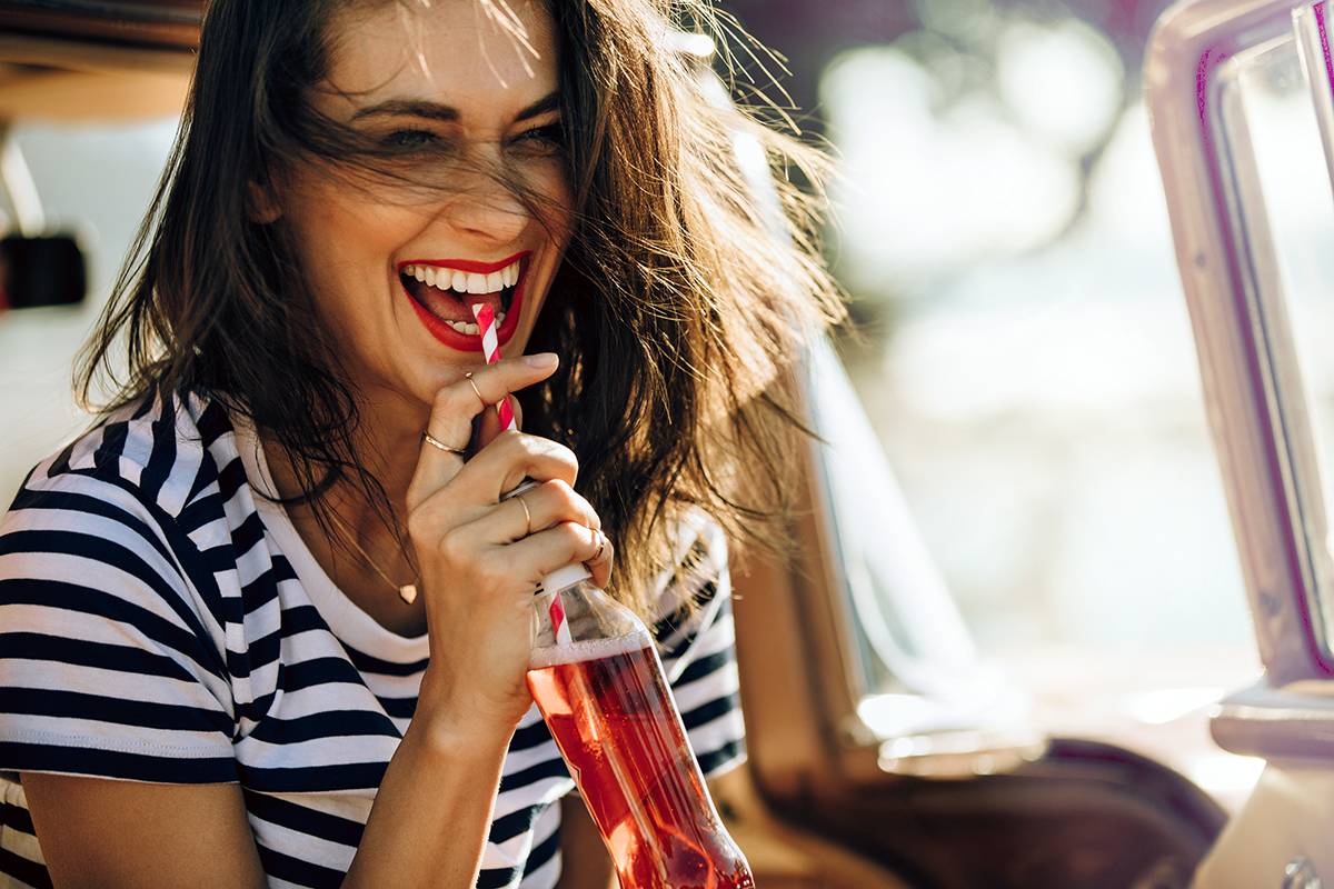 Closeup of beautiful young woman enjoying drinking cola with straw while sitting in a car. Female on a road trip having soft drink.