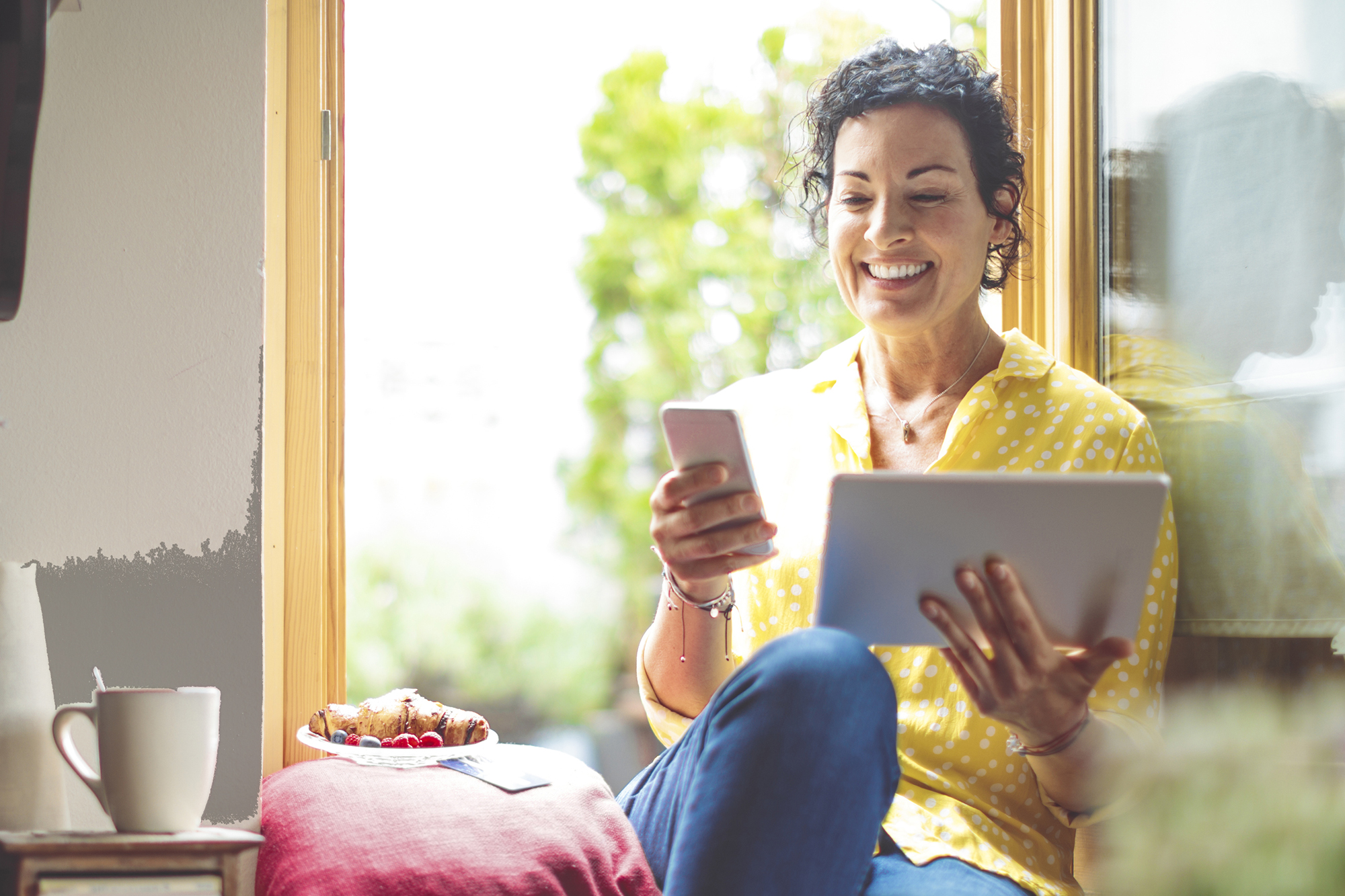 Mature woman is purchasing online at home Mature woman is purchasing online at home - using mobile phone and tablet - while sitting in yellow window, wearing yellow shirt, with a plate of pastry and coffee mug