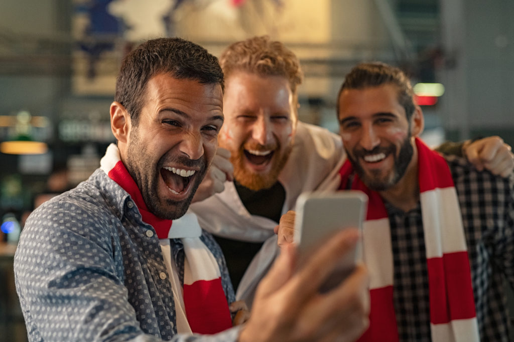 Excited men watching football in streaming on smartphone in bar. Football fans watching game on smart phone and celebrating victory score at pub. Happy supporters cheering and exulting after winning an online bet.
