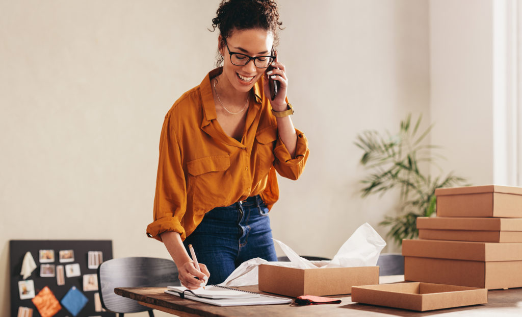 Drop shipping business owner talking on mobile phone and taking order. Female entrepreneur working at home office confirming the order on phone.