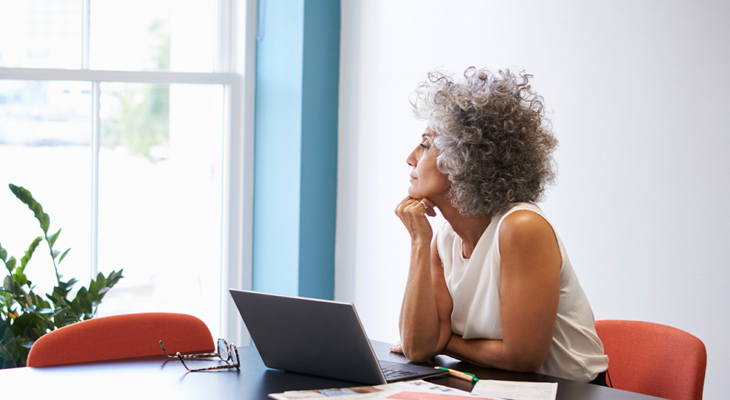 senior female thinking while looking out window, leaning on desk with open laptop and paperwork on desk
