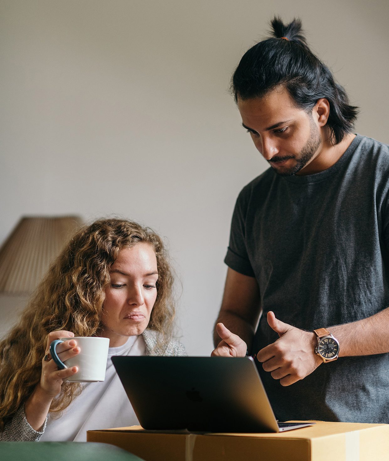 male with two thumbs up and female holding coffee mug, looking at laptop set up on a cardboard box