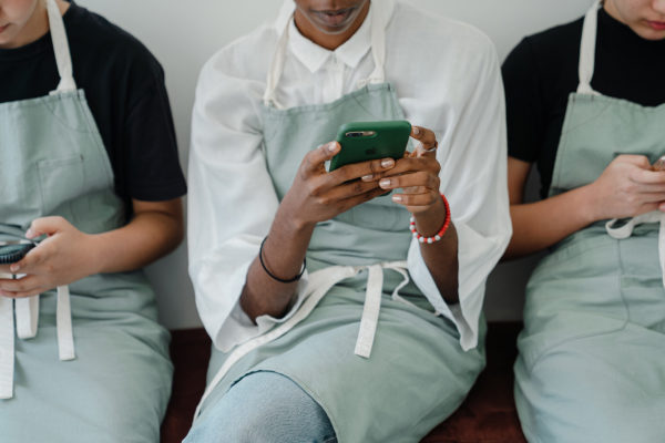 three adults wearing light green aprons, sit in a row, staring down while using their mobile phones