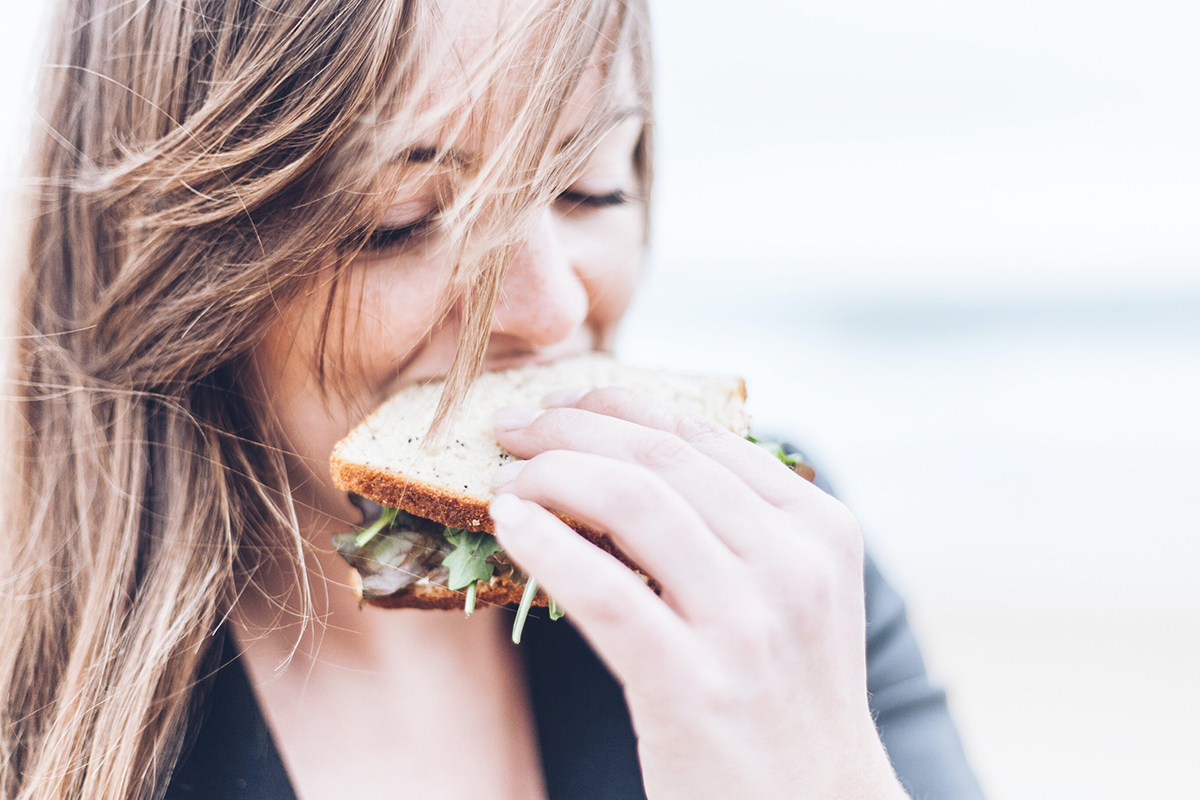 close up of woman eating sandwich with lettuce and deli meat, with lots of light in the background