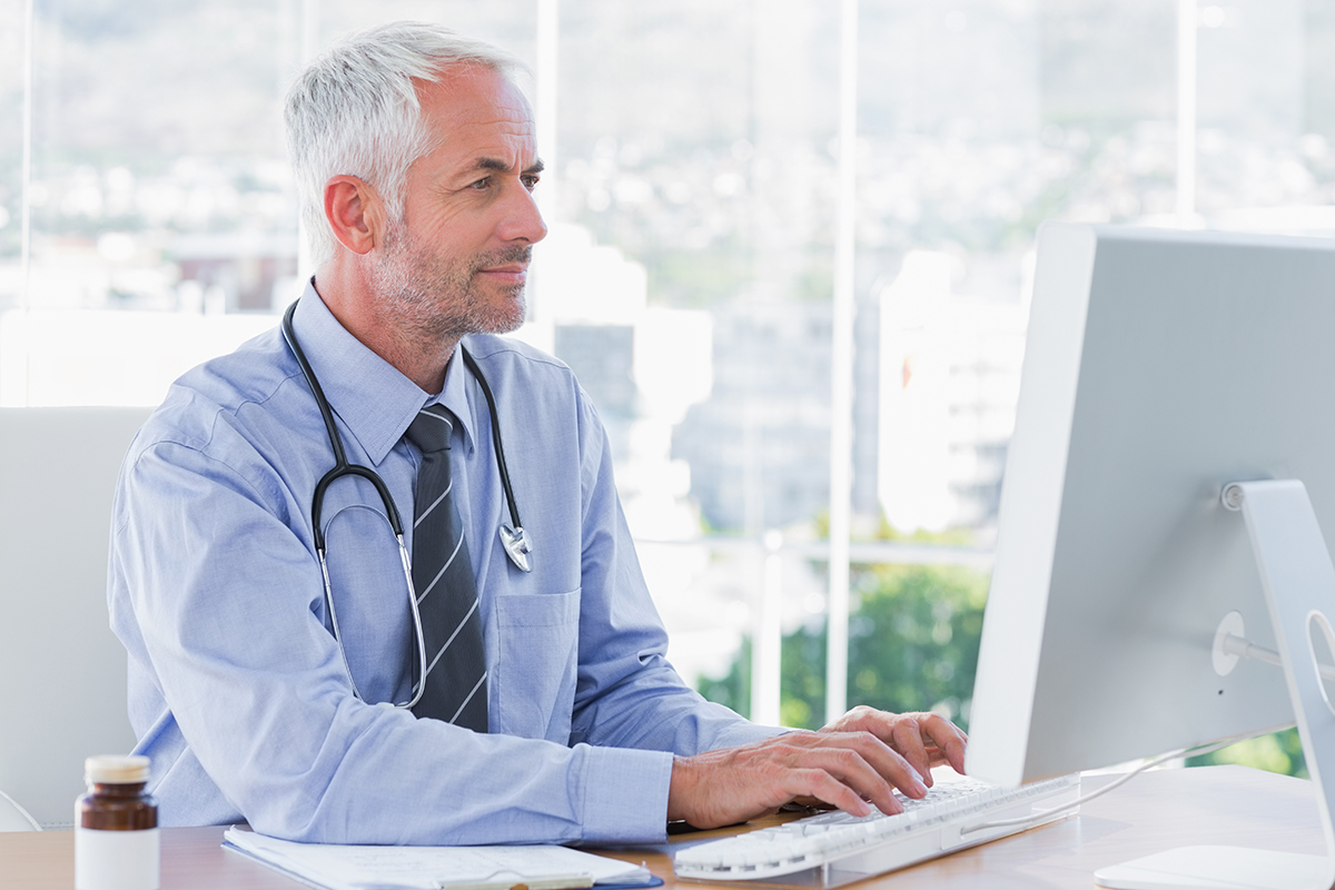 Older male doctor using desktop computer in bright office, wearing stethoscope, lots of light coming in windows