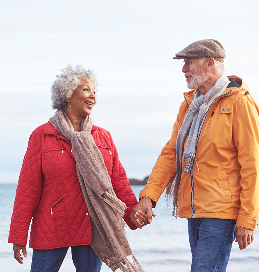 two seniors walking on the beach with the ocean in the background, slightly blurry, they are holding hands and looking at each other, smiling, wearing wamr jackets and scarves