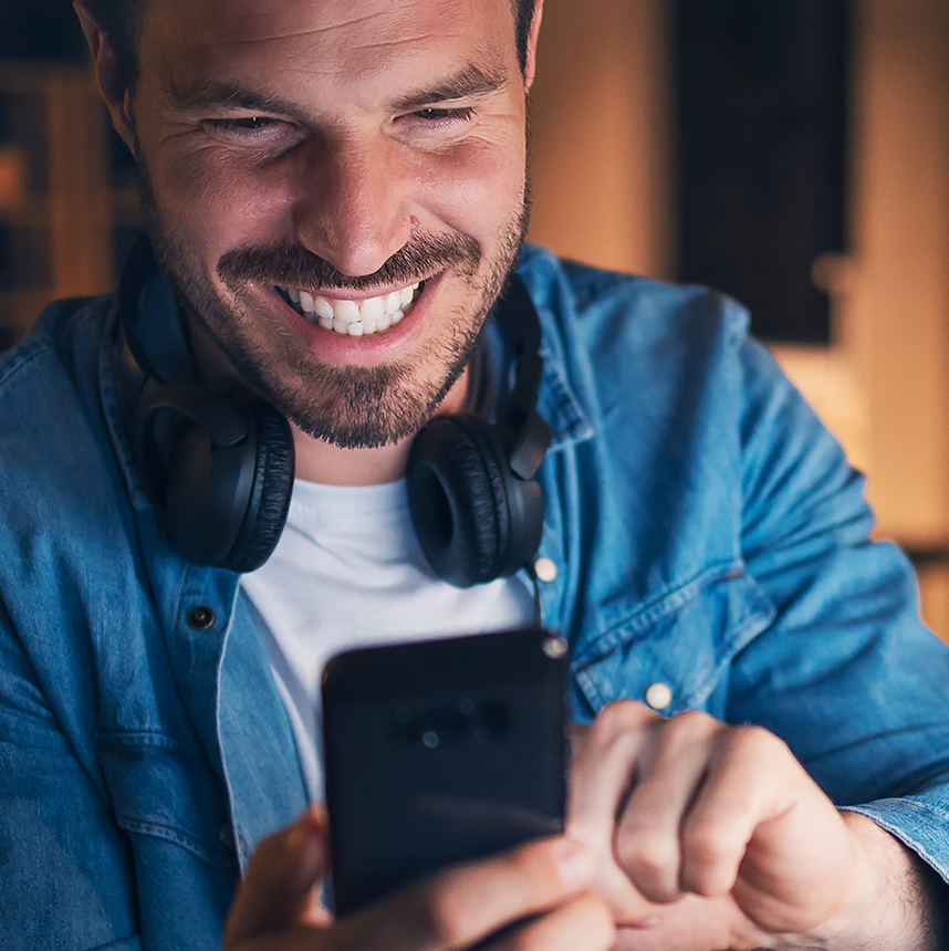 Male in blue shirt and wearing headphone around his neck, smiling while using mobile for gaming or sport betting