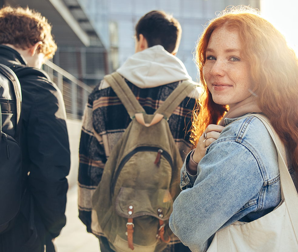 three students with backpacks, two males seen from behind, slightly blurry background, one female smiling and turning to face the camera in front