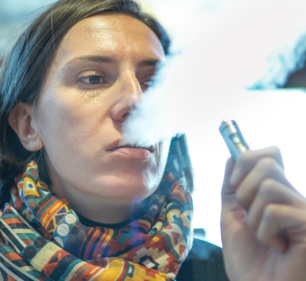 female vaping, she is wearing multicoloured scarf and her face is half covered by vape smoke coming out of her mouth while she exhales